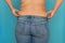 Back view of unrecognizable fat plump woman standing in blue jeans, squeezing excess naked waist on blue background.