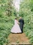 Back view of the walking newlyweds along the road covered with green bushes.