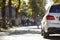 Back view of white car parked on city pedestrian zone on background of blurred silhouettes of people walking along green sunny