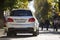 Back view of white car parked on city pedestrian zone on background of blurred silhouettes of people walking along green sunny