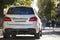 Back view of white car parked on city pedestrian zone on background of blurred silhouettes of people walking along green sunny