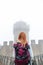 Back view woman with backpack wearing jeans jacket. Fortification, old castle on background. Journey. Vertical photo. Mystical