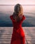 Back view of a woman in a luxurious red dress stands near the river. Atmospheric sunset on the pier.