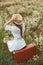 back view of woman in white dress with bouquet of wild camomile flowers sitting on retro suitcase