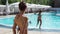 Back view yong slim woman in bikini, sunglasses and hat standing near poolside and looking at pool
