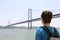 Back view of young backpacker man looking at bridge. Traveler or tourist with backpack on the waterfront in Lisbon Portugal next