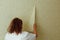 Back view of young fat woman wearing white T-shirt, beige trousers, standing at wall, removing old yellow wallpaper.