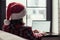 Back view of young freelancer woman in a red santa claus christmas hat sitting near window and working on laptop. Home work