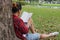 Back view of young relaxed man in red shirt leaning against a tree and reading textbook in beautiful outdoor park.