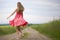 Back view of young romantic slim woman in red dress with long hair walking by ground road along green field on sunny summer day on