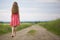 Back view of young romantic slim woman in red dress with long hair walking by ground road along green field on sunny summer day on