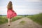 Back view of young romantic slim woman in red dress with long hair walking by ground road along green field on sunny summer day on