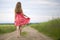 Back view of young romantic slim woman in red dress with long hair walking by ground road along green field on sunny summer day on