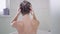 Back view of young slim woman shampooing hair sitting in bath at home. Brunette Caucasian lady washing head in slow