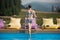 Back view young woman coming out from the water of a swimming pool at the resort, near stands a glass with a drink