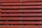 Back of a wooden bench, painted brown, consisting of horizontally fastened narrow slats, in the center attached to the metal base