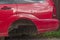 Back of wrecked red car that was damaged in accident, standing on wooden supports on grass near fence of brown metal profile