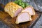 Backed Kasseler pork steak as piece and slice in puff pastry and egg yolks on a wooden cutting board with herbs
