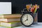 Background of sets of office items and a black alarm clock on colorful books, next to a glass with pencils, there is a place for