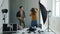 Backstage of studio photoshoot: guy posing for photographer while stylist moving clothing