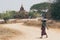 Bagan, Myanmar - March 2019: Shepherd grazing a gaunt cow through the dry field with temples and pagodas of ancient Bagan on