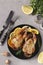 Baked quails with lemon and orange served on a dark plate on a gray background, Top view, Vertical orientation