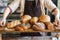 Baker in brown apron holding wooden tray with assorted freshly baked white flour, rhye, multigrain, wholegrain, challah breads, br