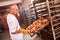 Baker holds a tray with fresh hot bread in his hands against background of shelves with pastries in bakery. Industrial production