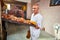 Baker holds a tray with fresh hot bread in his hands against background of shelves with pastries in bakery. Industrial production