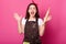 Baker young woman puts her hands up holding rolling pin, wears brown apron, white tshirt, opens her mouth widely. Adorable cute