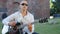 A bald adult man in sunglasses plays guitar and sings outdoors. Medium shot.