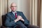 Bald attractive businessman in grey suit, blue shirt, red tie and glasses sitting in a chair in a hotel room.