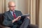 Bald attractive manager businessmen sitting on a chair in his hotel room looking at tablet.