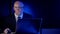 Bald businessman, in a jacket with a tie and glasses, middle-aged, sitting in the dark behind a laptop, looking at the