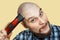 Bald guy with a beard and a clipper in his hands, shaving himself in the morning at home on a colored background