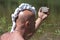 A bald guy with a shaving foam mohawk looks in the mirror. A man shaves in nature near a river with green grass. Ecotourism or