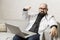 A bald male doctor in a white coat with glasses sits with a laptop on the couch. Online consultations. Quarantine during the