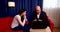 A bald man and a brunette with long hair are sitting in red chairs near a window with a blue curtain. the guy shows the