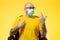 A bald man in medical mask with glasses sits in a chair and gestures emotionally. Yellow background. Concept of self-isolation,