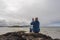 Bald man sitting on a rock by the ocean looking at his smart phone. Beautiful nature scenery in the background. West coast of