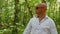 A bald man in sunglasses sings and walks through the woods. Green trees, summer, nature. Medium shot.