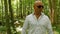 A bald man in sunglasses sings and walks through the woods. Green trees, summer, nature. Medium shot.