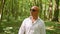 A bald man in sunglasses sings and walks through the woods. Green trees, summer, nature. Medium shot.