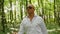 A bald man in sunglasses sings and walks through the woods. Green trees, summer, nature. Medium shot.