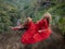 Bali trend photo. Caucasian woman in long red dress sitting on big stone in tropical rainforest. Vacation in Asia. Travel