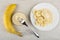 Banana, piece of banana strung on fork above bowl with condensed milk, plate with slices of banana, milk on table. Top view