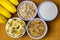 Banana sliced ramekin with oatmeal, granola and plain yogurt as side dishes under bamboo mat with bunch of bananas besides