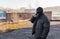 A bandit in a black leather jacket and a mask talking on the phone on the street near an abandoned building