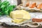 A bar of butter is cut into pieces on a wooden board with a knife, surrounded by milk, eggs and parsley on a brown table. Ingredie