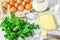 A bar of butter is cut into pieces on a wooden board with a knife, surrounded by milk, eggs and parsley on a white table. Ingredie